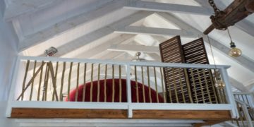 Cozy loft with a wooden railing overlooks a room, featuring a large, curved red sofa and a slatted wooden partition. Exposed beams add rustic charm.