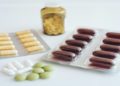 Various capsules and tablets are arranged on a white surface, including brown capsules in blister packs, yellow pills in a jar, and loose white and green tablets.