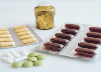 Various capsules and tablets are arranged on a white surface, including brown capsules in blister packs, yellow pills in a jar, and loose white and green tablets.