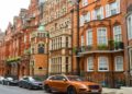 Street view of ornate red-brick Victorian townhouses in a row, lined with parked cars. The scene feels elegant and historical with lush greenery.