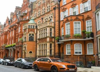 Street view of ornate red-brick Victorian townhouses in a row, lined with parked cars. The scene feels elegant and historical with lush greenery.