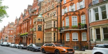 Street view of ornate red-brick Victorian townhouses in a row, lined with parked cars. The scene feels elegant and historical with lush greenery.