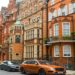 Street view of ornate red-brick Victorian townhouses in a row, lined with parked cars. The scene feels elegant and historical with lush greenery.