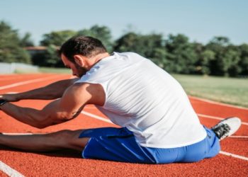 Man stretches on a red running track, wearing a white shirt and blue shorts. Trees and a clear sky are in the background, conveying a calm, focused mood.