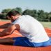 Man stretches on a red running track, wearing a white shirt and blue shorts. Trees and a clear sky are in the background, conveying a calm, focused mood.