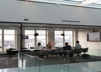 Four people are seated around a table in a modern glass-walled conference room, engaged in discussion. A presentation screen is visible. Bright and professional ambiance.