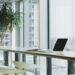 Modern office with large windows, a laptop on a sleek desk, surrounded by minimalist white chairs. A potted plant adds a touch of nature. Bright, airy ambiance.