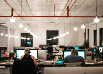 Modern open-plan office with people working at desks lined with computers. Bright round lights hang from the ceiling, creating a focused atmosphere.