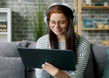 Woman wearing headphones and glasses sits on a gray couch, smiling at a tablet. She is in a cozy living room with a brick wall and shelves.