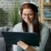 Woman wearing headphones and glasses sits on a gray couch, smiling at a tablet. She is in a cozy living room with a brick wall and shelves.