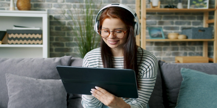 Woman wearing headphones and glasses sits on a gray couch, smiling at a tablet. She is in a cozy living room with a brick wall and shelves.