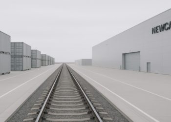 Railroad tracks lead to the horizon in an industrial area with stacked shipping containers and a large grey building labeled "NEWCASTLE" on a cloudy day.