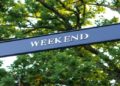 Black directional sign reads "Weekend" against a backdrop of lush green trees, creating a serene and inviting atmosphere.