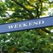 Black directional sign reads "Weekend" against a backdrop of lush green trees, creating a serene and inviting atmosphere.