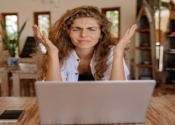Woman with long curly hair looks frustrated while using a laptop at a wooden table, surrounded by homey decor and a smartphone beside her.