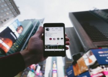 Hand holds a smartphone displaying a social media profile against the backdrop of tall, colorful, and lit-up city buildings, suggesting a lively urban setting.