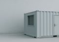 Minimalistic scene featuring a grey shipping container with a door and window, set against a plain grey wall. The tone is simple and industrial.