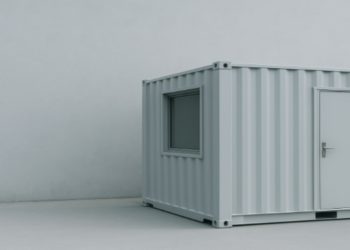 Minimalistic scene featuring a grey shipping container with a door and window, set against a plain grey wall. The tone is simple and industrial.
