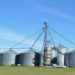 Row of large metal grain silos and an elevator stand on a flat grassy field under a clear blue sky, conveying an industrial yet peaceful rural scene.