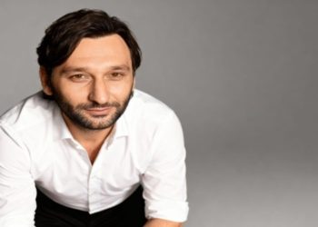 Andrei Marius Popescu with short dark hair and beard, wearing a white shirt, looking forward with a slight smile in a neutral gray background. The pose is relaxed and confident.
