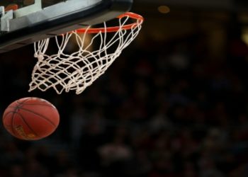 Basketball approaches a hoop, about to score, emphasizing action and precision in a dimly lit stadium filled with a blurred crowd.