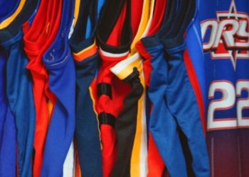 Row of colorful basketball jerseys hang closely together, featuring various designs and team colors. The scene conveys a sense of sport and team spirit.