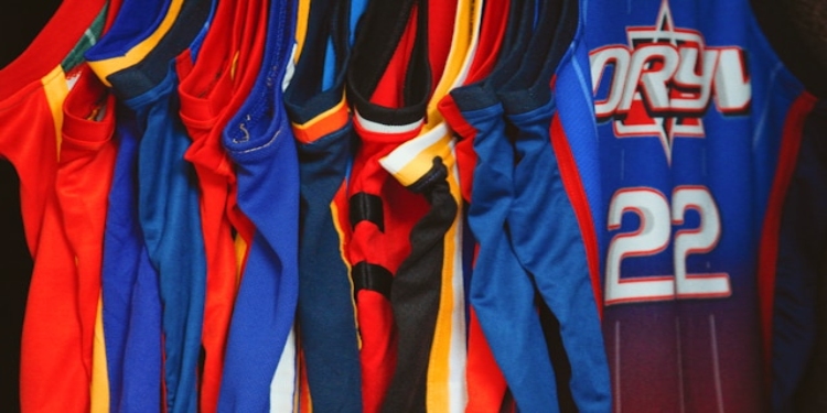 Row of colorful basketball jerseys hang closely together, featuring various designs and team colors. The scene conveys a sense of sport and team spirit.