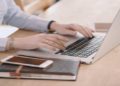 Person types on a laptop at a wooden desk, with a smartphone and notebook beside them. The scene conveys a focused, productive work atmosphere.
