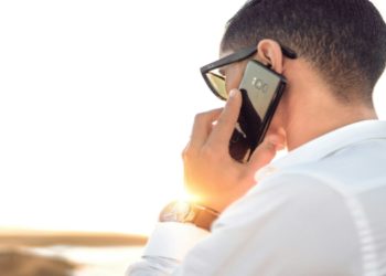 Person in sunglasses and a white shirt is talking on a smartphone outdoors at sunset. The scene conveys a calm and professional tone.