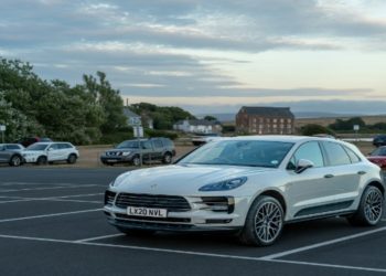 Silver car is parked in an almost empty lot under a cloudy sky at dusk, with a brick building and trees in the background, creating a serene scene.