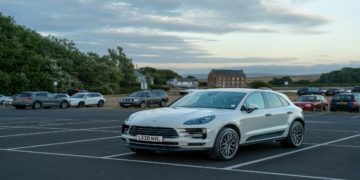 Silver car is parked in an almost empty lot under a cloudy sky at dusk, with a brick building and trees in the background, creating a serene scene.