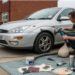 Man in a safety vest polishes a silver car on a residential street. Nearby, tools and car repair materials are laid out on a tarp, conveying a focused DIY atmosphere.