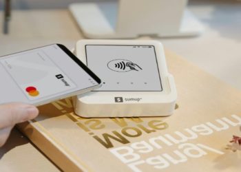 Hand holds a smartphone near a SumUp card reader, signaling a contactless payment. The setup rests on a desk with a calm, modern aesthetic.