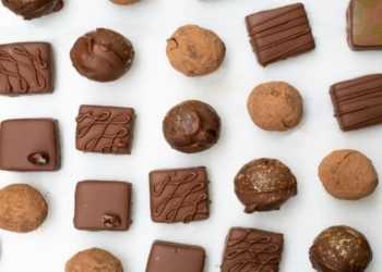 Assorted chocolates and truffles arranged in rows on a white background. The treats vary in shape and decoration, conveying a sense of indulgence.