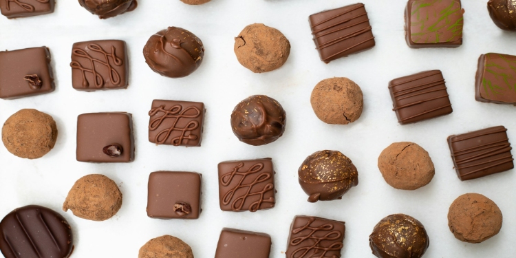 Assorted chocolates and truffles arranged in rows on a white background. The treats vary in shape and decoration, conveying a sense of indulgence.
