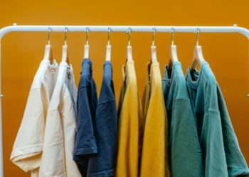 White clothing rack displays colorful T-shirts in shades of white, blue, yellow, and green against a vibrant orange background, creating a cheerful and stylish scene.