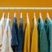 White clothing rack displays colorful T-shirts in shades of white, blue, yellow, and green against a vibrant orange background, creating a cheerful and stylish scene.