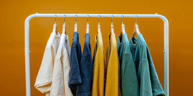 White clothing rack displays colorful T-shirts in shades of white, blue, yellow, and green against a vibrant orange background, creating a cheerful and stylish scene.