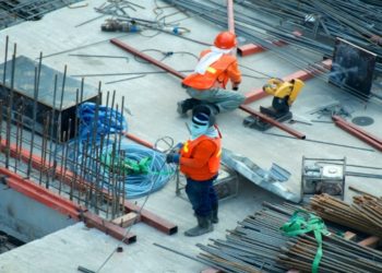 Two construction workers in orange safety gear and helmets stand on a concrete site with scattered metal rods and cables, conveying a busy, industrious environment.