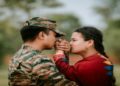 Soldier in camouflage uniform tenderly holds a woman's hand while she kisses it, both expressing affection outdoors with greenery in the background.