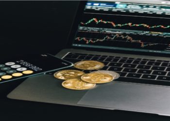 Laptop displaying stock charts, a smartphone with a calculator app showing numbers, and several gold bitcoins are placed on a dark surface, conveying a financial theme.