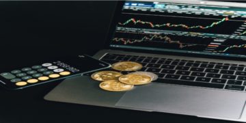 Laptop displaying stock charts, a smartphone with a calculator app showing numbers, and several gold bitcoins are placed on a dark surface, conveying a financial theme.