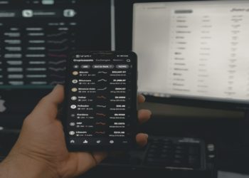A hand holds a smartphone displaying cryptocurrency prices against a dark background, with two computer monitors showing financial data. The scene conveys a high-tech, analytical atmosphere.