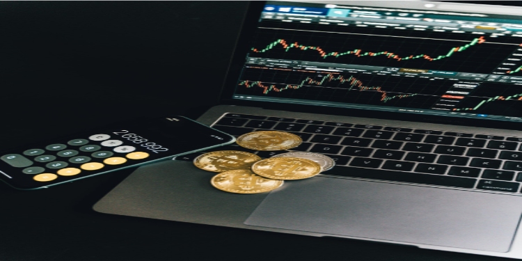 Laptop displaying stock charts, a smartphone with a calculator app showing numbers, and several gold bitcoins are placed on a dark surface, conveying a financial theme.