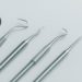 Five stainless steel dental tools, including a mirror and probes, are neatly arranged on a light gray background, conveying a clean, clinical atmosphere.