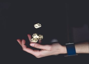 Hand skillfully juggling four white dice against a dark background, evoking a sense of focus and precision. The person wears a blue wristband.