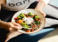 Person in workout attire holds a bowl of quinoa, broccoli, tomatoes, and dips, sitting on a yoga mat. The scene feels healthy and relaxed.