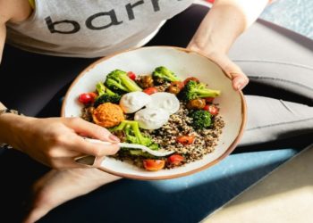 Person in workout attire holds a bowl of quinoa, broccoli, tomatoes, and dips, sitting on a yoga mat. The scene feels healthy and relaxed.