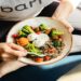 Person in workout attire holds a bowl of quinoa, broccoli, tomatoes, and dips, sitting on a yoga mat. The scene feels healthy and relaxed.