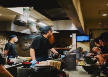 Japanese restaurant kitchen with chefs wearing black uniforms and hats preparing dishes. Diners seated at the counter enjoy their meals, creating a lively atmosphere.